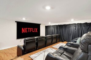 Une pièce dotée d’un grand écran affichant le logo « NETFLIX », de fauteuils en cuir inclinables, d’une table en bois et d’un rideau noir en arrière-plan.
