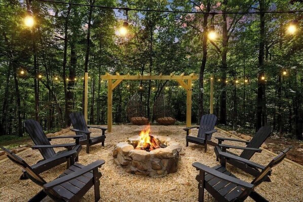 Un espace extérieur aménagé avec des chaises Adirondack, un foyer en pierre et des guirlandes lumineuses, le tout dans un cadre forestier.