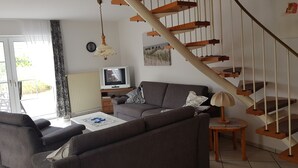 Un salon avec un canapé gris, un téléviseur posé sur un meuble en bois, une lampe et un escalier avec des marches en bois.