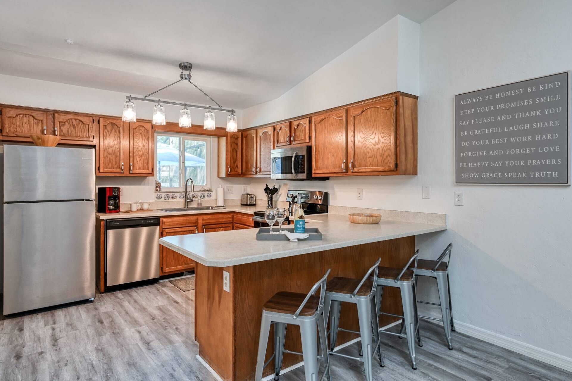 A modern kitchen with wooden cabinets, stainless steel appliances, a central island with bar stools, and a motivational quote on the wall.
