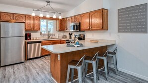 A modern kitchen with wooden cabinets, stainless steel appliances, a central island with bar stools, and a motivational quote on the wall. - River's Edge Retreat (St. Augustine)