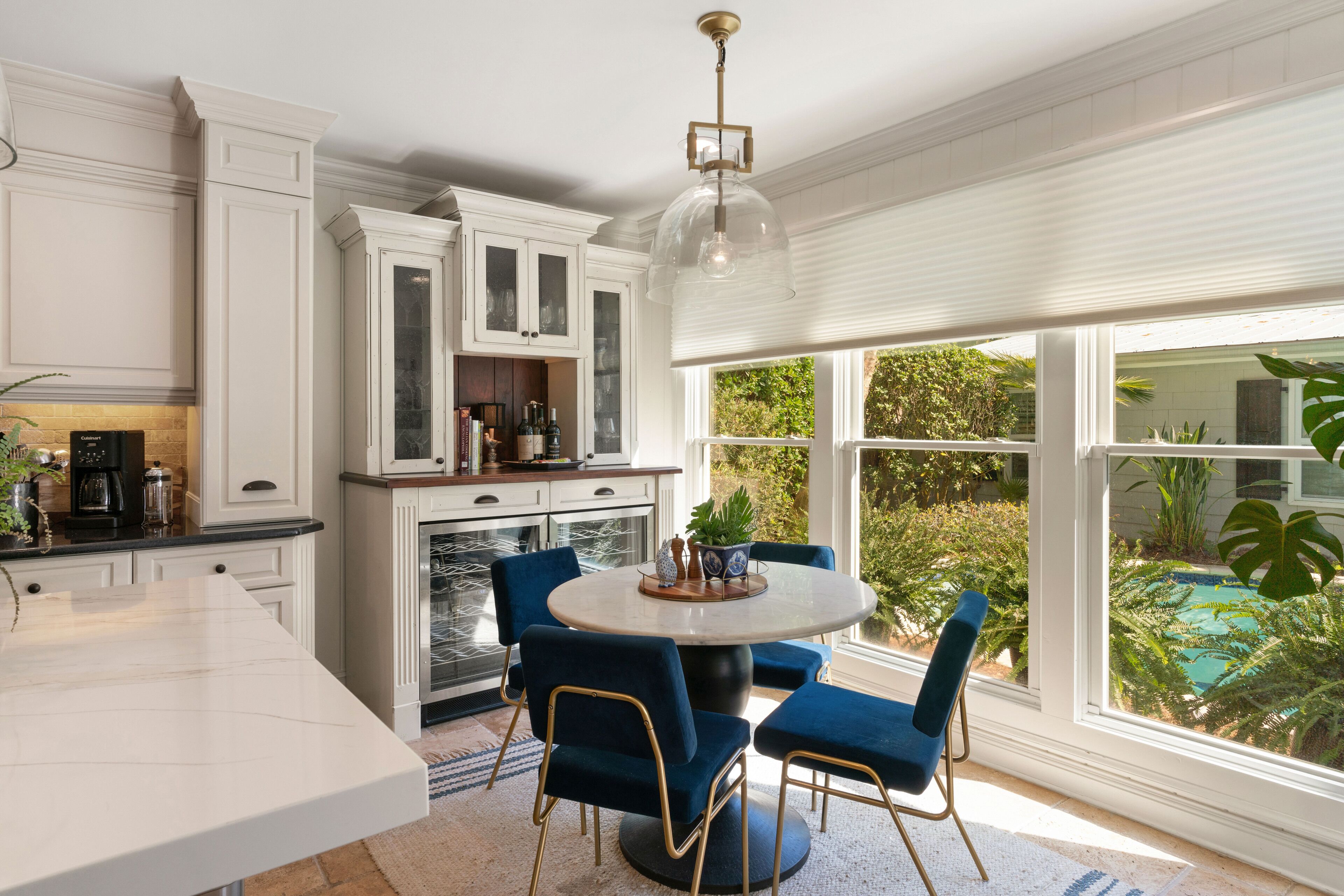 A kitchen with white cabinetry, a dining area with a round table and blue chairs, and a view of a swimming pool through large windows.