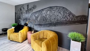 Une pièce avec deux fauteuils jaunes, une fresque murale grise représentant un pont, une petite table ronde et une plante en pot.