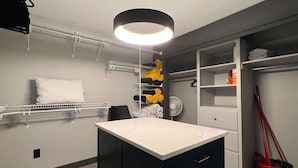 Une petite pièce comprenant une coiffeuse blanche, un meuble noir, une étagère blanche et un balai rouge.