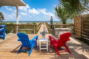 Eine Holzterrasse mit zwei Adirondack-Stühlen, einem kleinen Tisch und einem Whirlpool. Von der Terrasse aus hat man Blick auf einen Strand mit einem hölzernen Steg, der zum Meer führt.