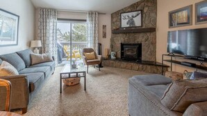 A living room with a stone fireplace, a sofa, a television, and a view of the outdoors. - Fraser 3BR Condo with Pool and Hot Tub (Fraser)