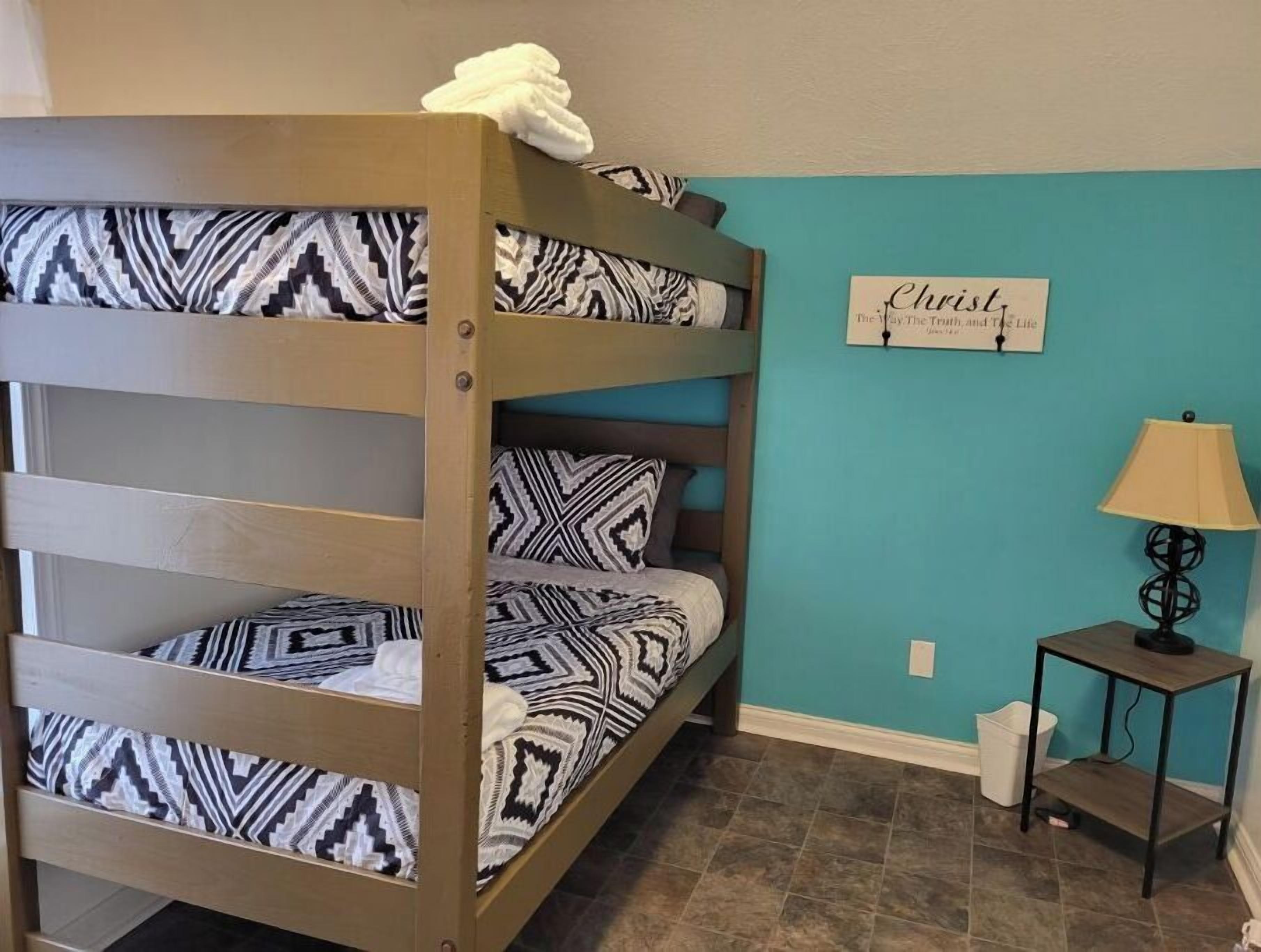 Una cama litera con ropa de cama estampada, una pared con una cita religiosa, una lámpara y un cubo de basura.