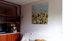 Een slaapkamer met een bed, een bloemschilderij aan de muur, een houten plank met een waterkoker en een plafondlamp.