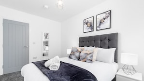 Ein modernes Schlafzimmer mit grauem, gepolstertem Kopfteil, weißer Bettwäsche und zwei gerahmten Kunstwerken über dem Bett.