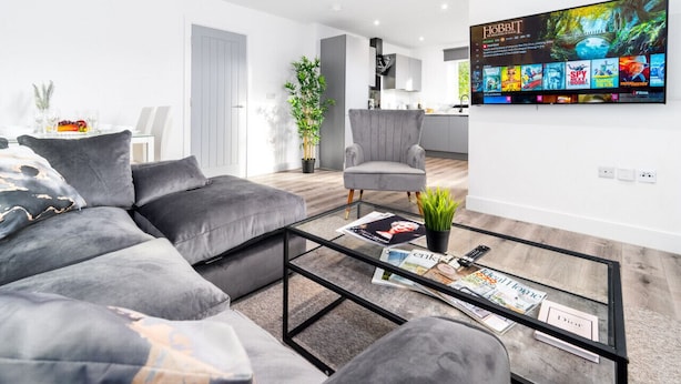 Ein modernes Wohnzimmer mit grauem Sofa, einem Glastisch und einem montierten Fernseher, der Filmempfehlungen anzeigt.