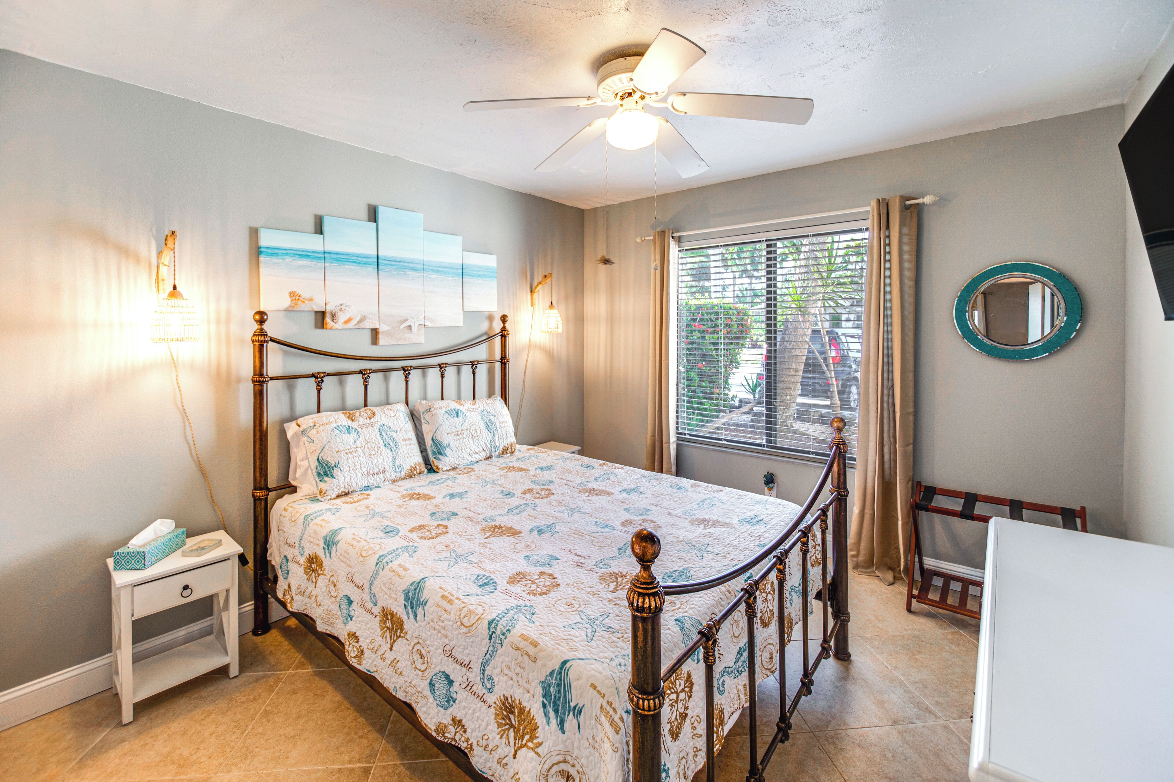 A bedroom with a bed, bedside table, nightstand, ceiling fan, window with curtains, and a wall-mounted mirror.