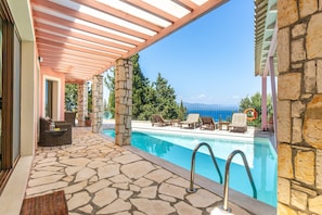 A covered outdoor pool area with stone flooring, lounge chairs, and a view of the sea.