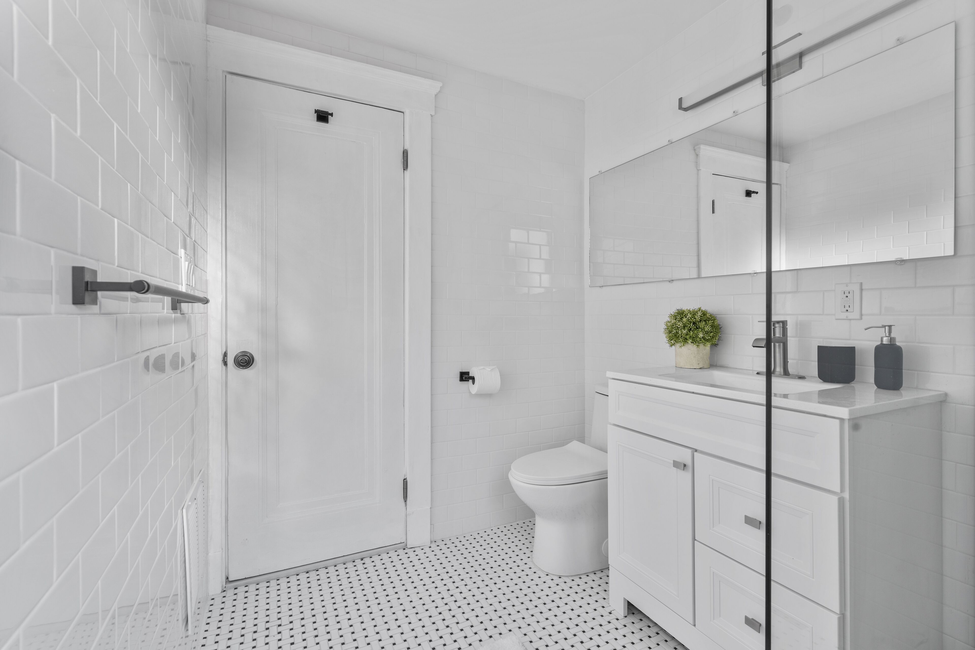 Un baño moderno con paredes de azulejos blancos, un inodoro blanco, cabina de ducha de vidrio y tocador blanco con espejo.