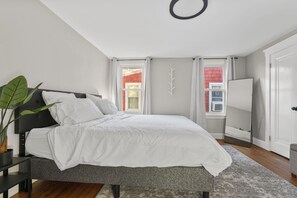Une chambre à coucher avec un lit, une table de chevet, une plante en pot, un miroir et une fenêtre avec des rideaux.