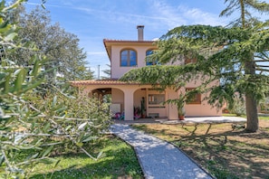 Un bâtiment à deux étages doté d’un patio couvert, d’un chemin de gravier menant à l’entrée et d’un grand arbre en premier plan.