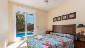 Une chambre à coucher comprenant un lit, une table de chevet, une lampe, un ventilateur de plafond, une porte-fenêtre coulissante donnant sur une piscine, et des tableaux encadrés accrochés au mur.