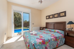 A bedroom with a bed, a nightstand, a lamp, a ceiling fan, a sliding glass door leading to a pool, and framed pictures on the wall.