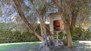 Une cabane perchée dans un arbre, dotée d’une structure en bois et d’un intérieur violet, entourée d’arbres et d’une zone herbeuse.