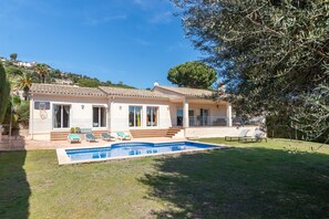 A two-story house with a swimming pool, a patio with sun loungers, and a view of a hill with houses.