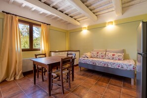 A room with a wooden dining table, two chairs, a sofa with floral bedding, a window with curtains, and a ceiling with exposed beams.