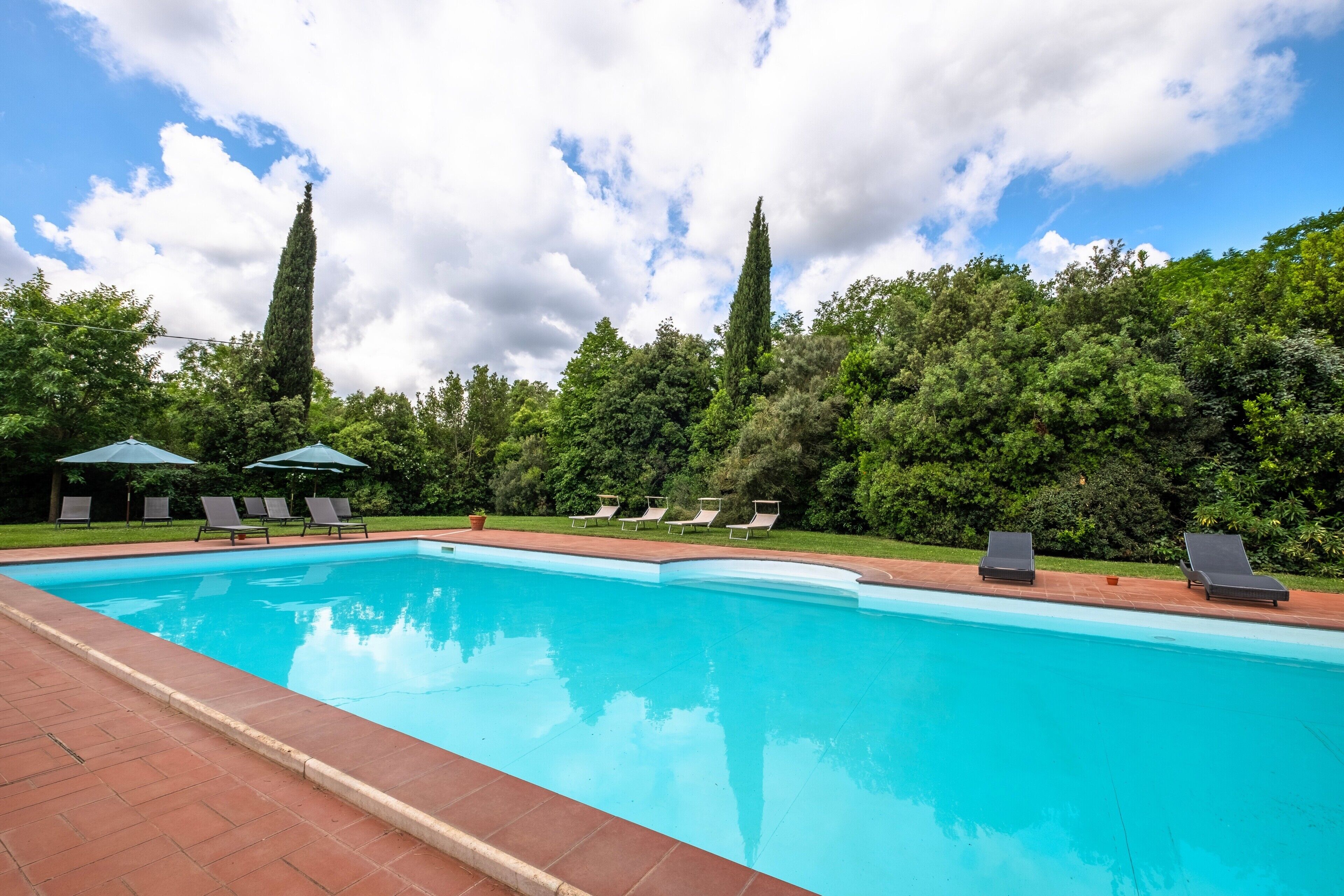 Una piscina con sillas de descanso y sombrillas, rodeada de árboles y un cielo despejado.