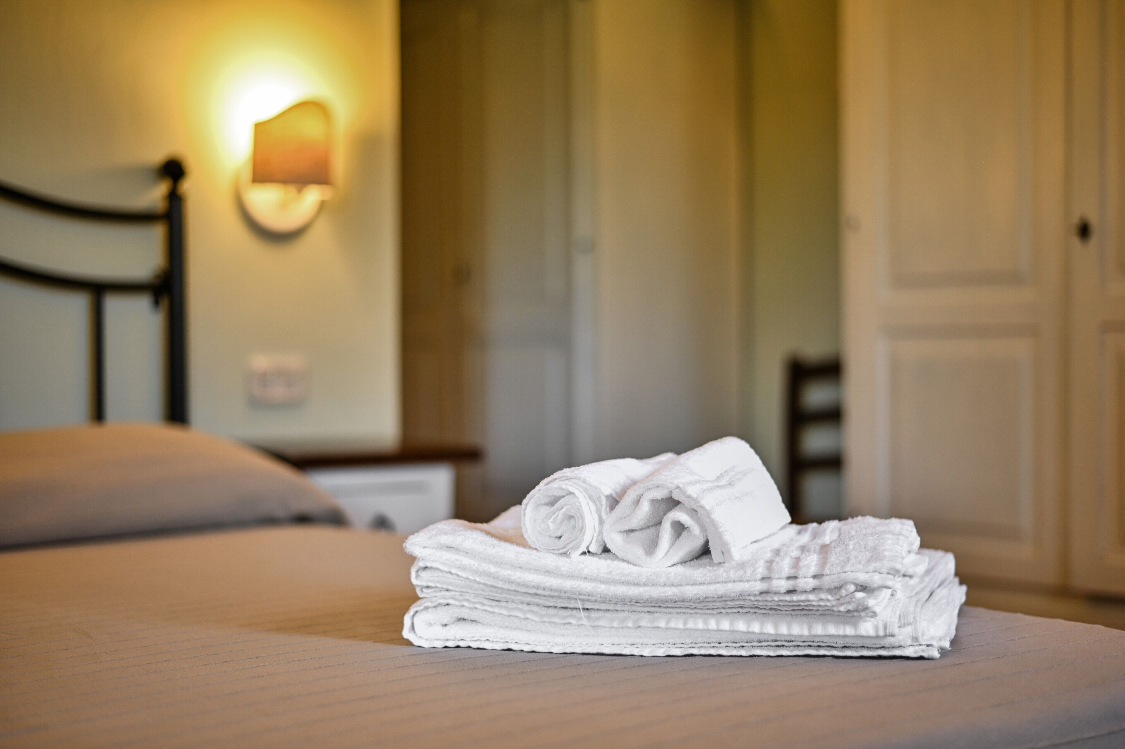 A neatly made bed with a folded white towel on top, a wall-mounted light fixture, and a door in the background.