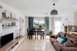 A living room with a TV, dining table, chairs, a brown sofa, and a chandelier.