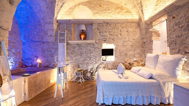 Een kamer met stenen muren, een bed met witte lakens, een tafeltje met een lamp en een witte stoel.