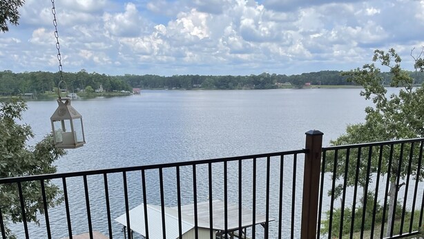 A wooden deck with two Adirondack chairs, a hanging lantern, and a view of a lake with a dock and pier.