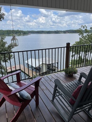 Eine Holzterrasse mit zwei Adirondack-Stühlen, einem hängenden Laternenleuchter und Blick auf einen See mit Steg und Pier.