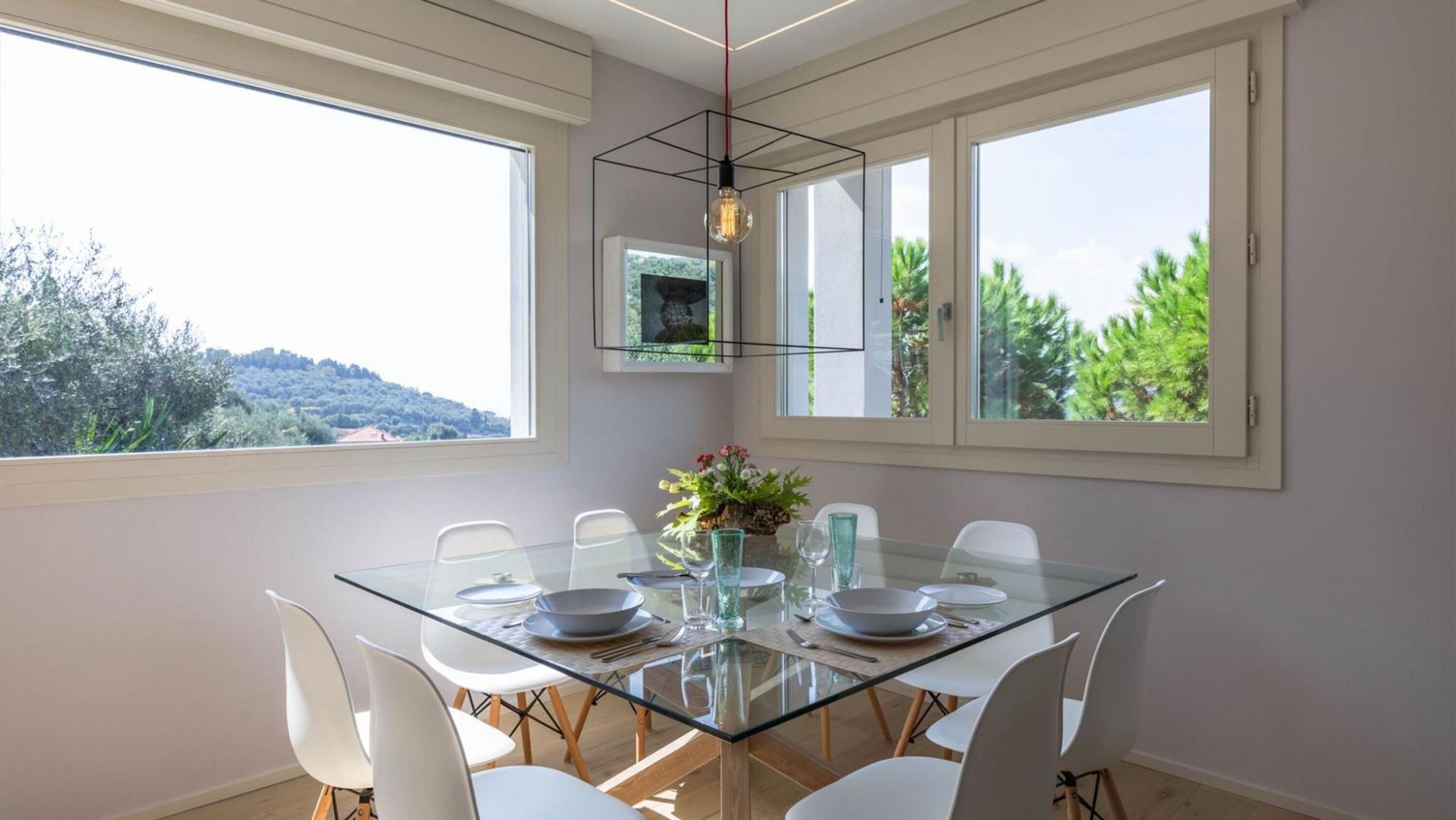 A dining area with a glass table, white chairs, and a view of greenery outside.