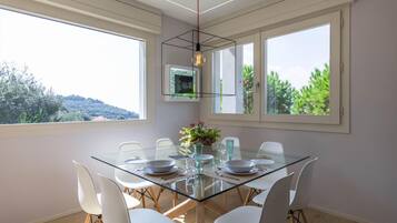 A dining area with a glass table, white chairs, and a view of greenery outside.