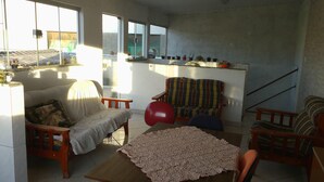 Una sala de estar con un sofá, sillones, una mesa con mantel de encaje y una silla roja. Hay plantas en macetas en un estante y una escalera al fondo.