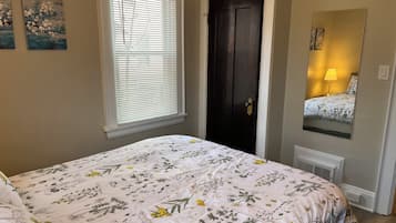 Ein Schlafzimmer mit blumigem Bettbezug, einem Fenster mit Jalousien, einer dunklen Tür und einem Spiegel, der ein Bett mit einer Lampe reflektiert.