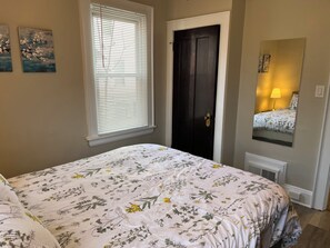 Ein Schlafzimmer mit blumigem Bettbezug, einem Fenster mit Jalousien, einer dunklen Tür und einem Spiegel, der ein Bett mit einer Lampe reflektiert.