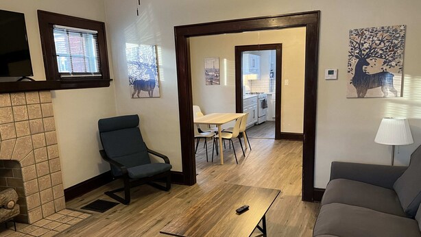 Un salon comprenant un canapé, un fauteuil, une table basse en bois et un coin repas avec une table et des chaises. On y trouve également une cheminée, un tableau et une lampe sur une table d’appoint.