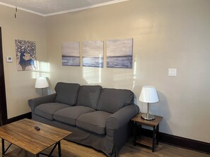 Ein Wohnzimmer mit grauem Sofa, einem Holztisch und zwei Stehlampen. An den Wänden hängen drei Landschaftsbilder.
