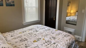 Une chambre à coucher avec une couvre-lit à motifs floraux, une fenêtre avec des stores, une porte sombre et un miroir qui reflète un lit avec une lampe.