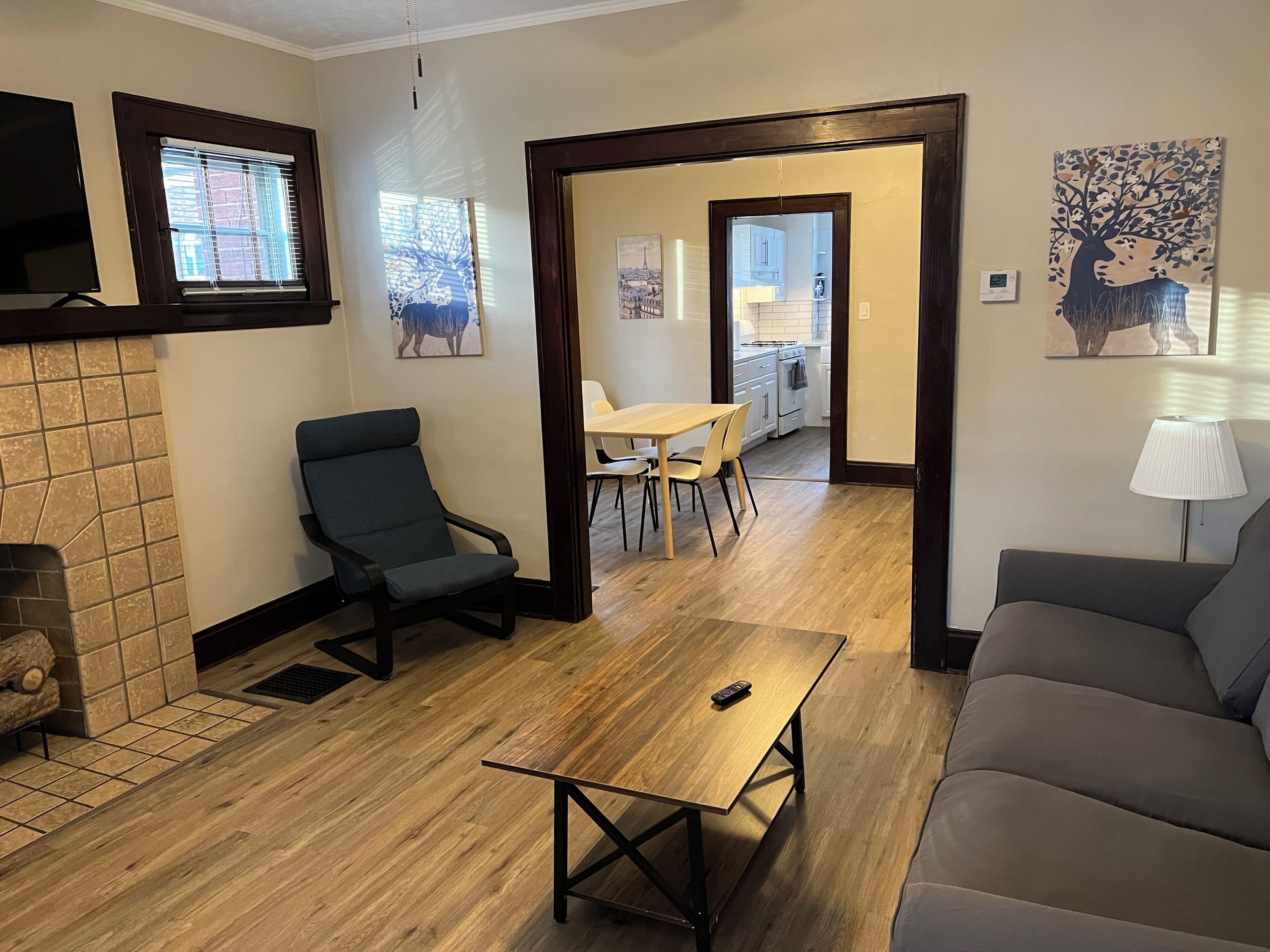 Un salon comprenant un canapé, un fauteuil, une table basse en bois et un coin repas avec une table et des chaises. On y trouve également une cheminée, un tableau et une lampe sur une table d’appoint.