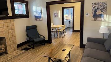 Un salon comprenant un canapé, un fauteuil, une table basse en bois et un coin repas avec une table et des chaises. On y trouve également une cheminée, un tableau et une lampe sur une table d’appoint.