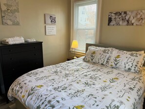 A bedroom with a floral bedspread, a dresser with towels, and wall art including a quote and a floral painting.
