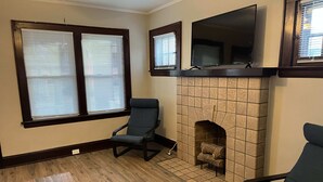 Un salon avec une cheminée, deux fenêtres équipées de stores, un téléviseur fixé au-dessus de la cheminée et deux fauteuils.