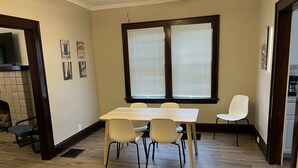 Un coin repas comprenant une table en bois et quatre chaises, une cheminée et une grande fenêtre équipée de stores.