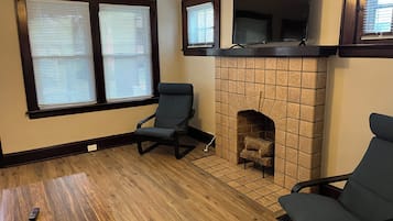 Ein Wohnzimmer mit Kamin, zwei Fenstern mit Jalousien, einem Fernseher über dem Kamin und zwei Sesseln.