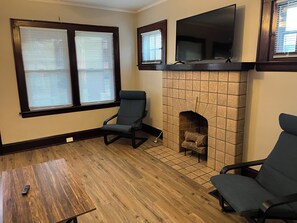 A living room with a fireplace, two windows with blinds, a TV mounted above the fireplace, and two chairs.