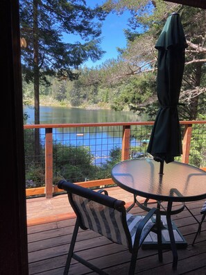 A wooden deck with a glass table and chairs, overlooking a calm lake surrounded by trees. - Lakeside cabin on the Central Oregon Coast  (Florence)