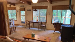 Ein gemütliches Wohnzimmer mit einer Couch, einem Holztisch und einem Kamin. Es gibt einen Essbereich mit Tisch und Stühlen, sowie große Fenster mit Jalousien.