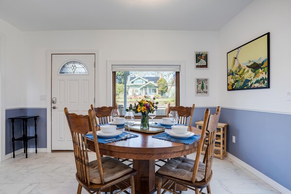 A dining room with a round wooden table set for six, surrounded by wooden chairs with cushioned seats. A vase with flowers is placed in the center of the table.