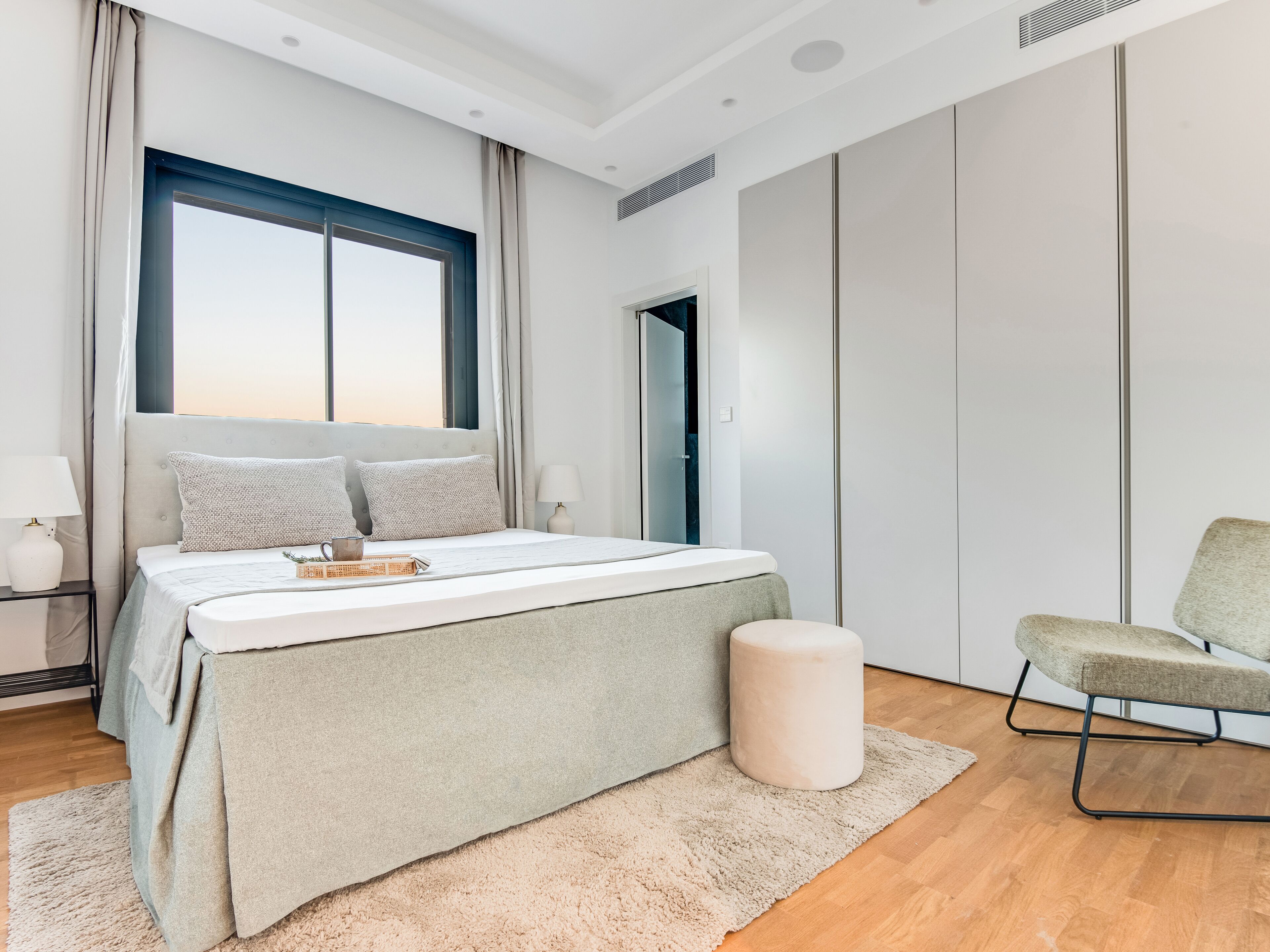 A modern bedroom with a large bed, a bedside table, a chair, a window, and a wardrobe.