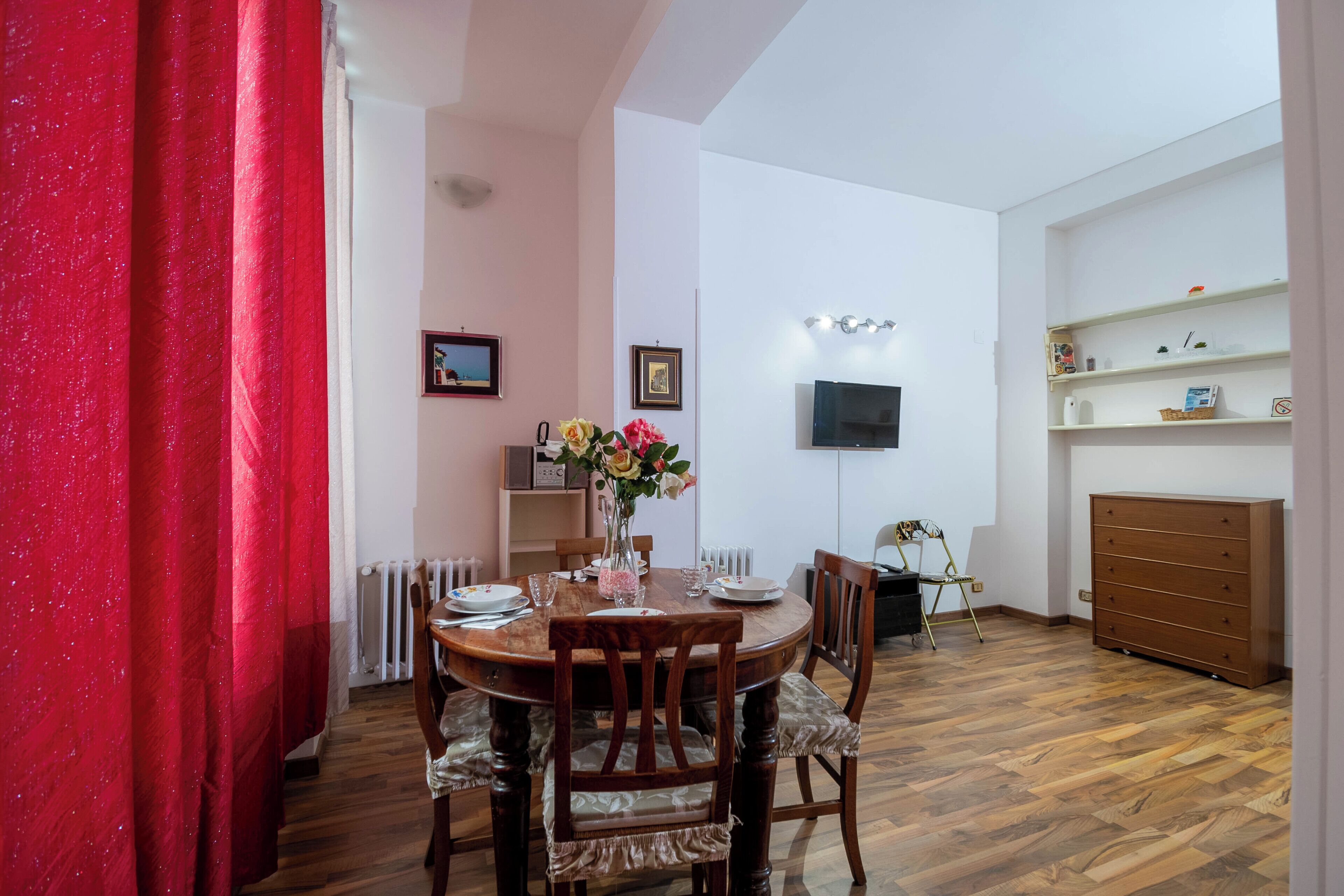 A dining area with a round table, chairs, a vase with flowers, a television on the wall, and a wooden dresser.